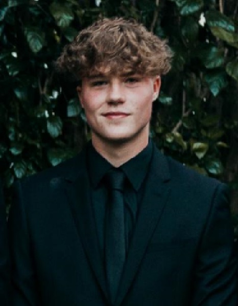 Head and shoulders shot of a young man standing in front of green shubbery He has a mop of brown curly hair up to his ears, and he is smiling. He is wearing a dark shirt, tie and jacket.
