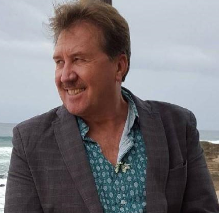 Head and shoulders shot of a middle aged man with short brown hair. He is looking to the side, smiling. He stands in front of the ocean and is wearing an open neck blue shirt with a grey jacket