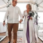 Wedding picture of counsellor Kate Crosson and her husband, on a white background, showing him in brown long pants and a white shirt, wearing open sandles. She is wearing a long flowing white robe. In her one hand, she is carrying a bouquet of flowers, and she is holding his other hand, while they are walking down a wooden floor, looking at each other. He is wearing sun glasses and has short brown hair. Her hair is long flowing golden locks.