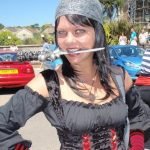 Head and shoulders shot of Counsellor Kate Crosson in her younger years, SHe has straight black hair and is wearing a beanie. Her off the shoulders black top has sleeves up to her elbows, and shes wearing open finger gloves which have red and black stripes. She has a play play knife between her teeth and stands with her hands on her hips. In the background are cars, people and buildings. A blue sky and sunny day
