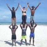 Six people on the beach sand with the ocean behind them, and blue skies on a sunny day. Three of these people appear quite young, and are holding onto the legs of the other three people who are standing on the shoulders of the first three people. Those standing on their shoulders, have their arms spread open, the outer two people are holding the hands of the person in the middle and their free hands are stretched towards the sky. The middle person standing on shuoulders, is Counsellor Kate Crosson. SHe is wearing black pants and a black bikini top. On either side of her, are dark skinned males. The one to her left, is wearing blue denim knee high shorts. The other, is wearing artificial leopard skin long pants, and a black top.