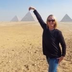 Kate Crosson standing on the desert sand facing us. She is wearing blue denim jeans and a loose fitting black top, and sunshades. Her blond hair is neatly cropped. She is smiling. Her one hand is raised to point down on a pyramid, which is in the middle of two others, in the distance. Blue sky and very sunny. Shadows from her legs are cast to the left of the picture