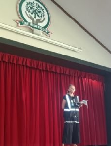 Kate Crosson stands on stage with a red curtain behind her. SHe is wearing a loose fitting dark pants and top, and is holding papers in her hand. Her blond hair is tied back. SHe is facing the audience.