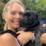 Head and shoulders shot of a blond haired lady wearing a light blue shirt is holding a black dog, looks like a Pug, in front of large trees,