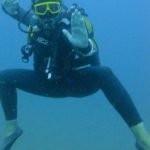 Frontal shot of a lady swimming beneath the water wearing a full body wet suit, flippers and yellow goggles. She is in tadpole stance with her one hand open outwards facing the viewer.