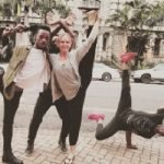 Standing on the pavement next to a street are four people in strange poses, of which three are Negro males and Kate Crosson. They are dressed in comfortable black pants and loose fitting shirts