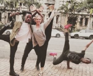 Standing on the pavement next to a street are four people in strange poses, of which three are Negro males and Kate Crosson. They are dressed in comfortable black pants and loose fitting shirts