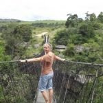 This picture shows a lady walking across a draw bridge over a gorge between hills which have huge rocks and trees among wild grass. She is turning back towards the camera, one hand on each of the rails, and is wearing denim knee high shorts and a light brown shirt. She has blond hair tied back on her head and is smiling